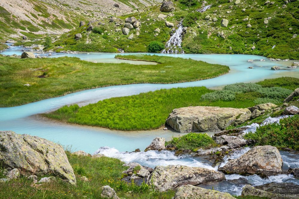 A glacier-fed brilliant turquoise creek meanders through a meadow with several waterfalls in Écrins National Park in France.