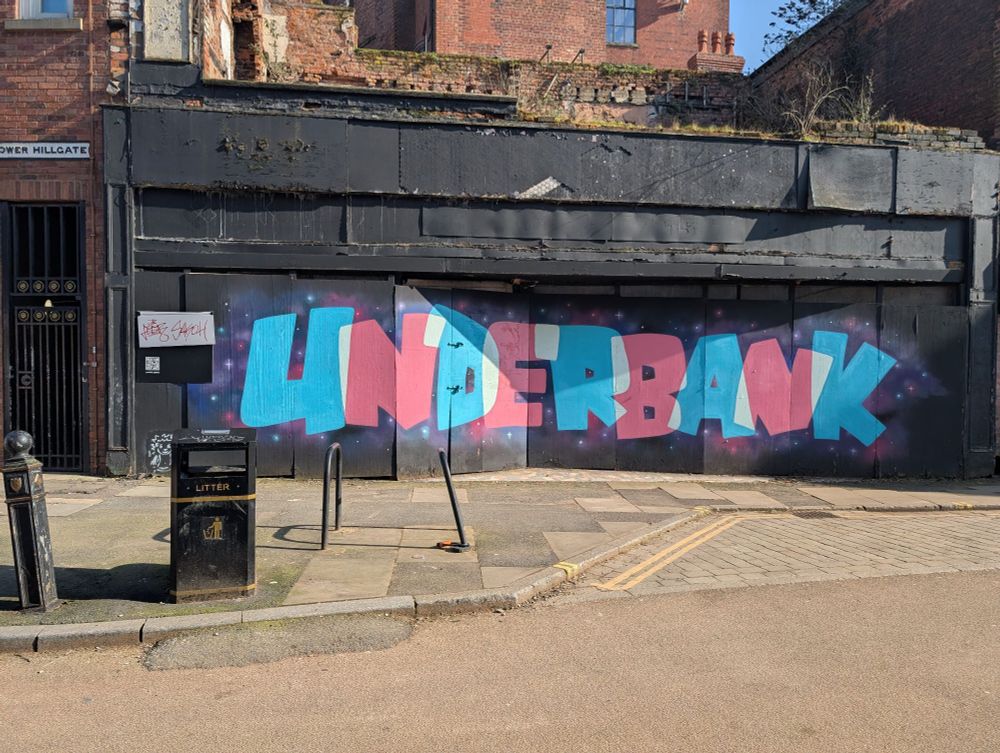 Bottom of Lower Hillgate, polite street art with "Underbank" spray painted in cyan and pink bubble all caps. There's a scrawled tag next to this, two cycle parking bars, one a bit bent over. 