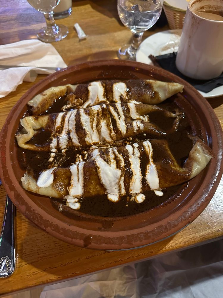 A photograph showing a table at a restaurant. A heavy, dark red ceramic bowl is filled with Crepas Con Mole- three large crepes filled with chicken and covered with a thick mole sauce, with white queso drizzled over the top. There are water glasses, a mug, silverware, and napkins on the wooden table in the background.