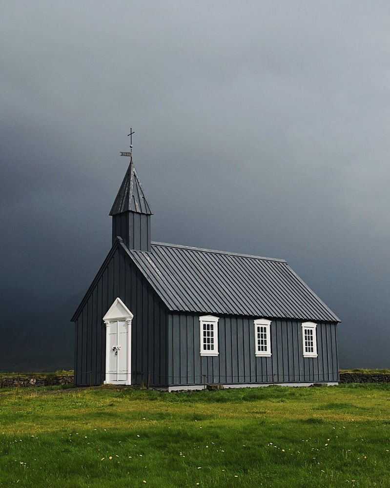 Grey wooden Icelandic church with white trim, against a moody sky with grass in the foreground.