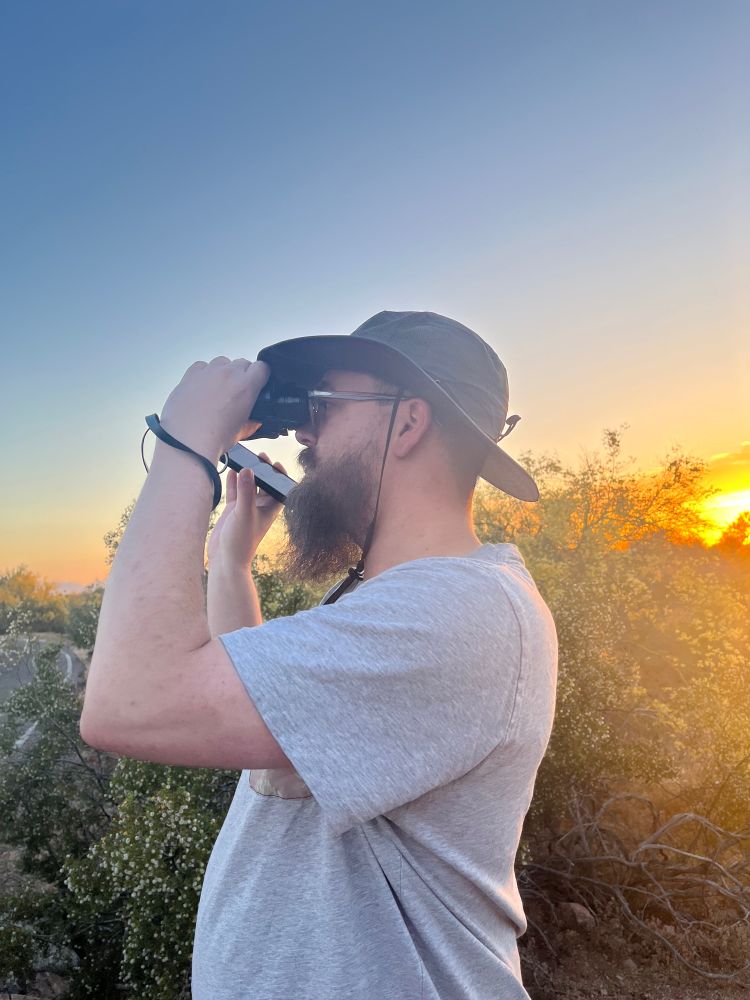 A lovely bearded man is looking off in the distance with a pair of binoculars at sunset