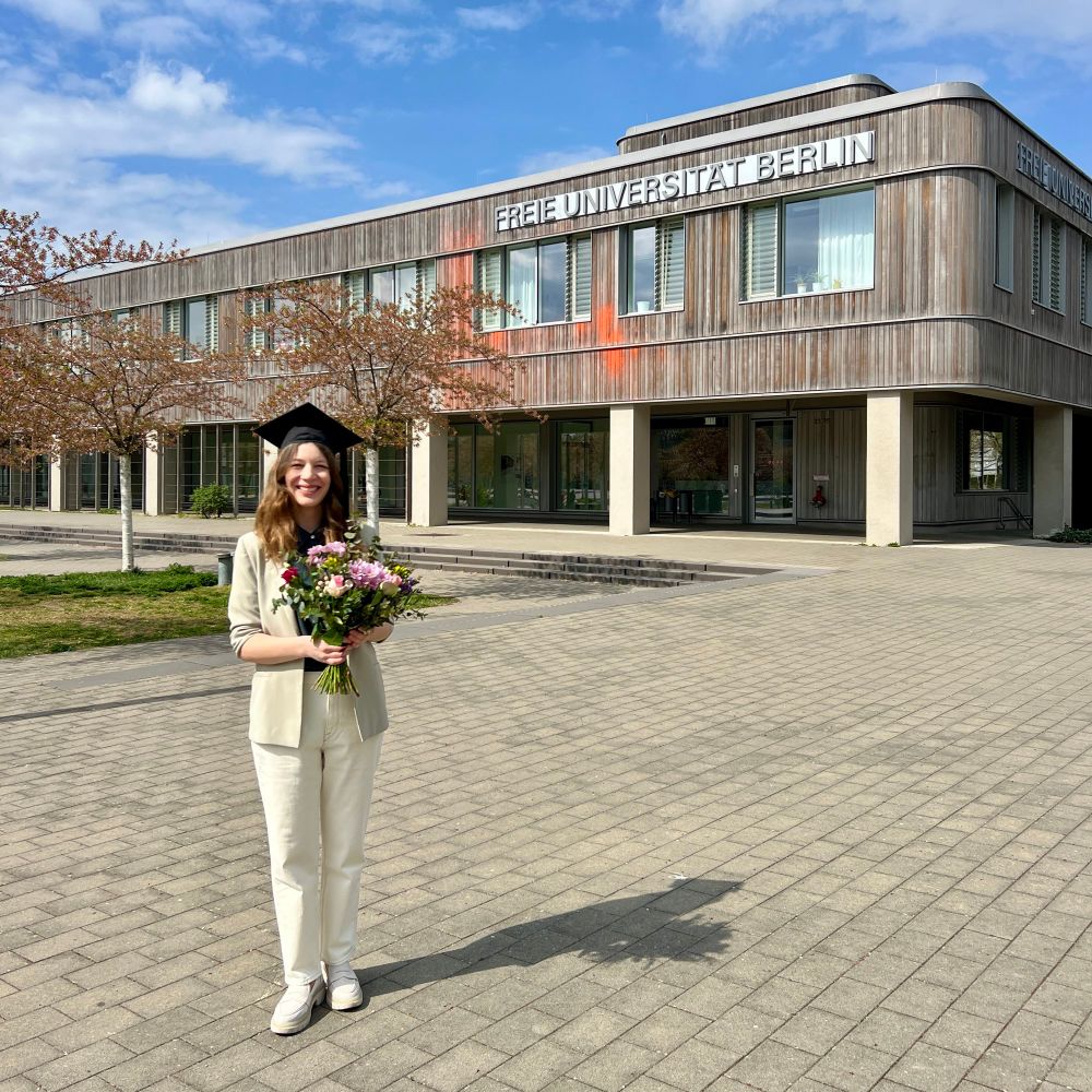 Woman wearing a graduation hat and holding flowers standing in front of Freie Universität Berlin