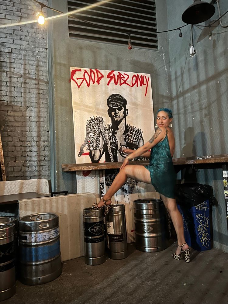 Mae poses in an over the shoulder smolder and one leg up in front on mural. The mural says good subs only featuring a leather daddy. 