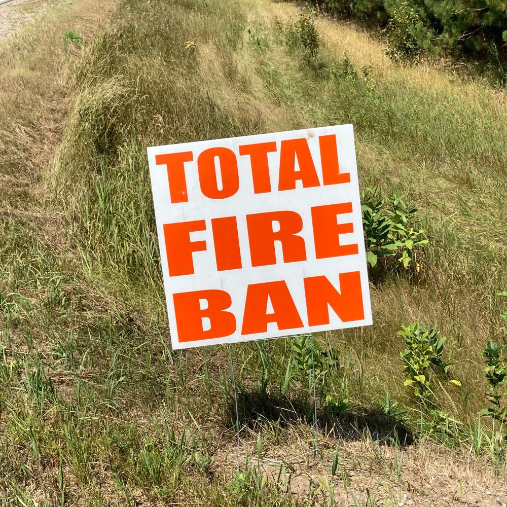 An orange and white road sign sitting at the edge of a road, surrounded by very dry grass. The text reads: Total Fire Ban.