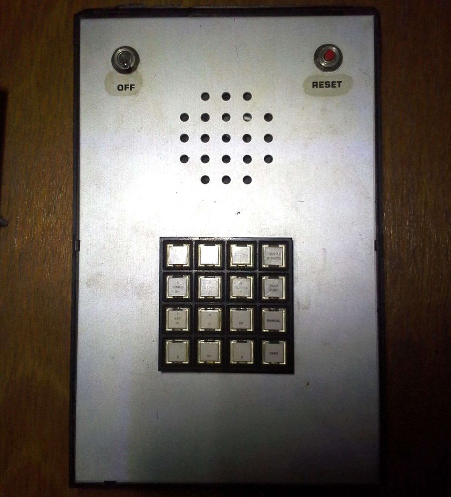 A metal plate on a black plastic box roughly the same size, a 16-key keypad below some holes for a speaker grille, both roughly centered on the plate, an on/off toggle switch in the upper left, and a red pushbutton in the upper right labeled "RESET".