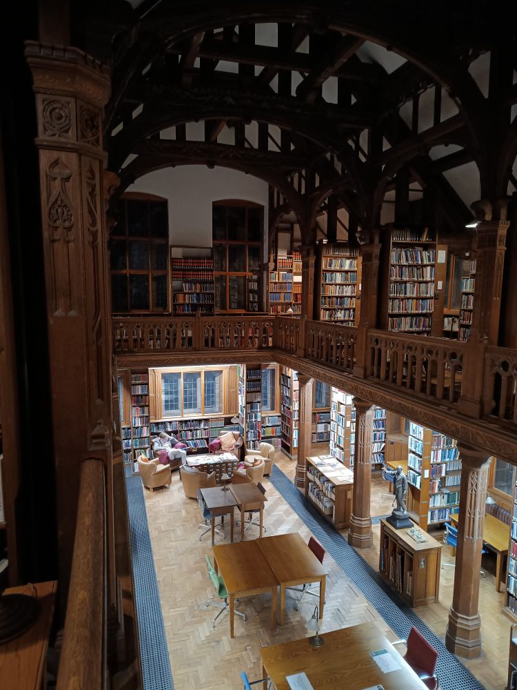 Photo of a room filled with books and with wooden pillars, beams all around. It's taken from the first floor looking down onto tables, a parquet floor. There are leaded windows but it's dark outside 