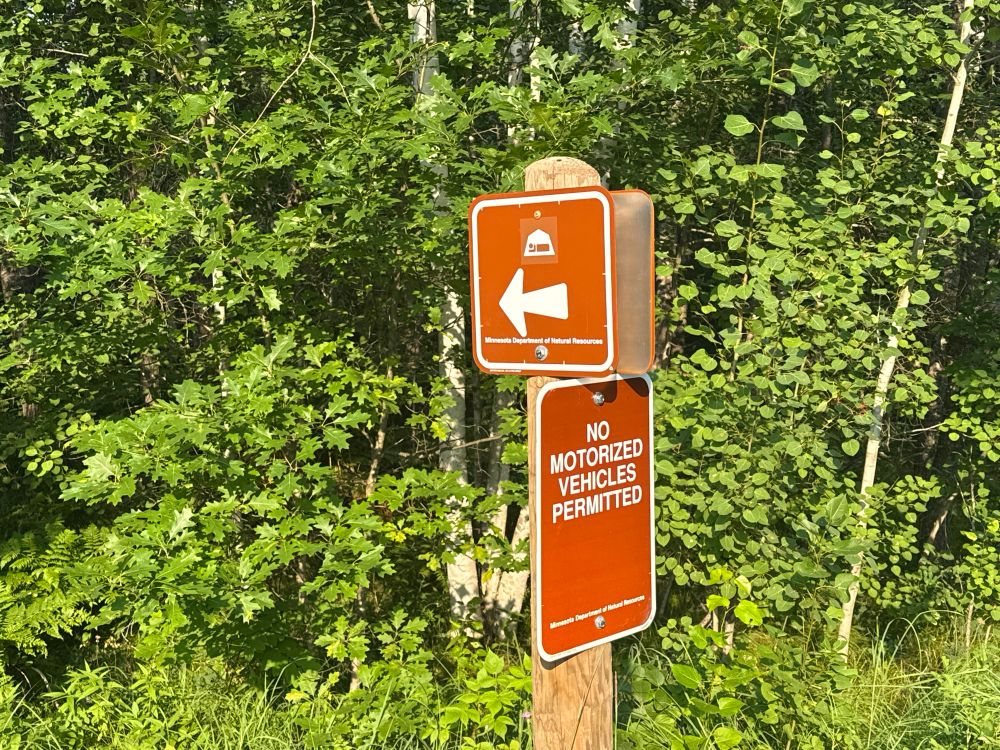 Photograph of 2 brown signs on a signpost in front of green trees. One shows a tent and an arrow indicating the direction to a camp site. The other says “No motorized vehicles permitted”