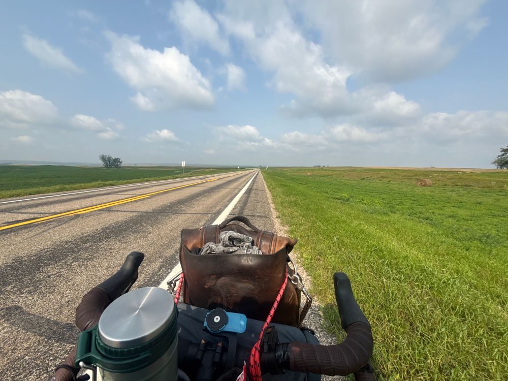 Photograph over bicycle handlebars showing a grey paved road stretching off into the relatively flat rolling hills of the North Dakota countryside. White clouds against a pale blue sky stretch to the hazy horizon 