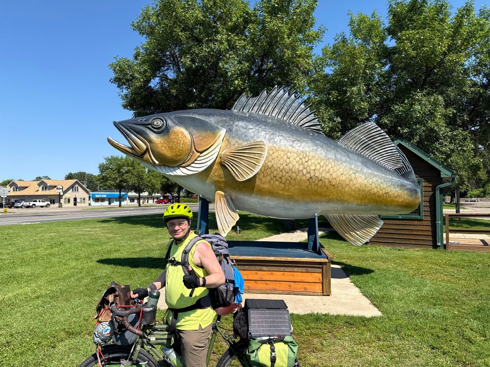 Photograph of me on my fully geared up touring bicycle n front of a statue of a big fish several times my size. 
