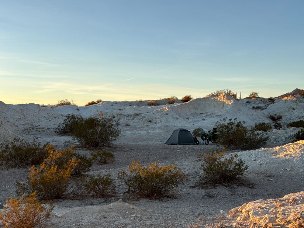 Photograph of a grey tent and a bicycle next to some creosote bushes in a small valley of white rocky ground under a blue evening sky. 