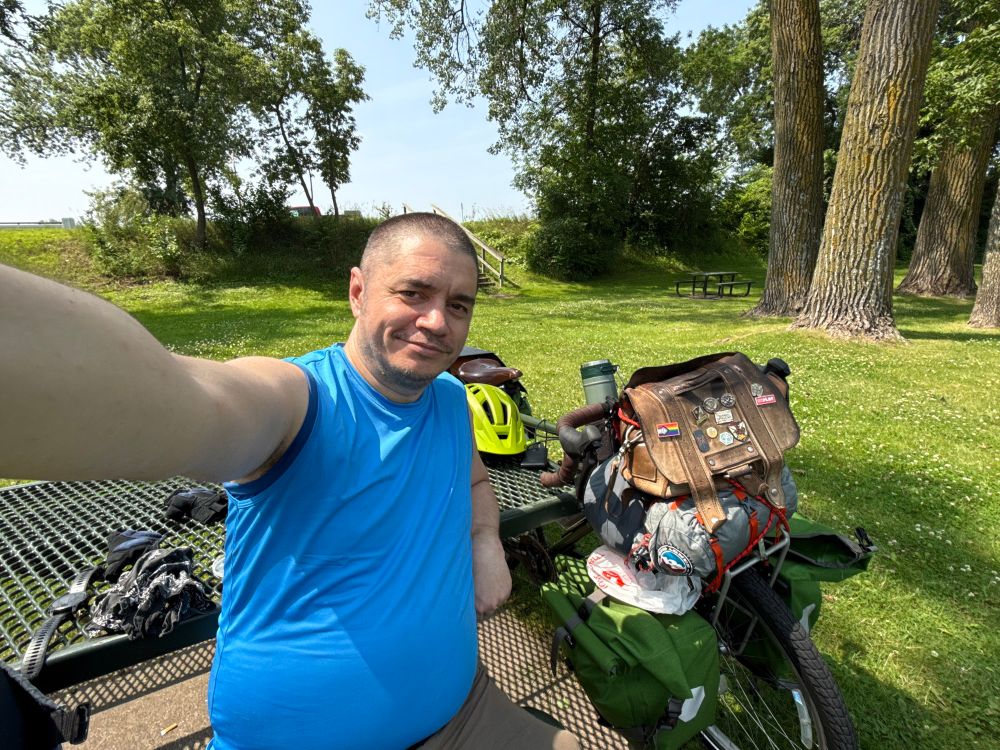 Selfie of me with buzzed hair wearing an aqua blue tank top sitting at a picnic table against which leans my heavily loaded Surly Long Haul Trucker touring bicycle. 