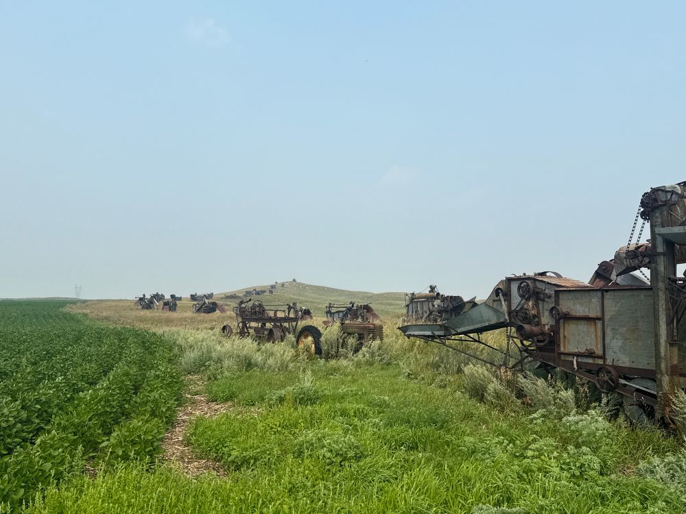 Photograph of a line of derelict threshing machines stretching along a ridge line between a dark green cultivated field on the left and lighter green prairie plants on the right.