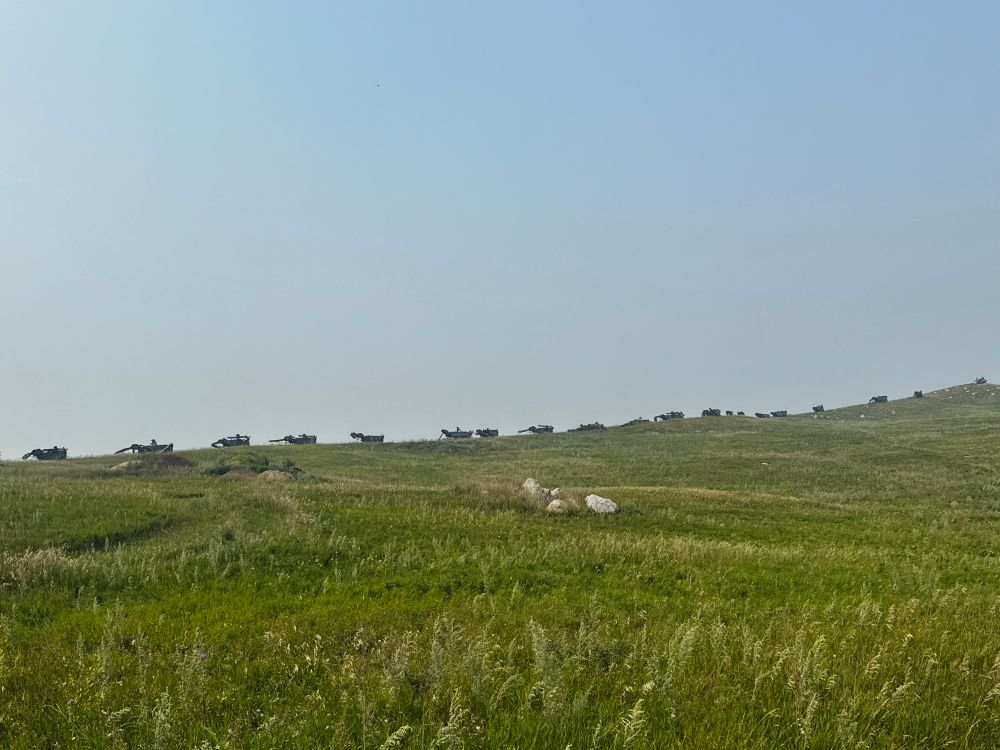 Photograph showing a line of derelict farm machinery along a ridge silhouetted against a pale blue sky with green grassland in the foreground 
