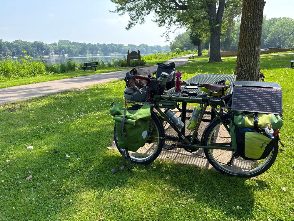 Photograph of a heavily loaded green Surly Long Haul Trucker bicycle leaning against a picnic table in green grass of a park by the Mississippi River. The bicycle has greens front and rear panniers, as well as sleeping gear and a camera bag on the front rack & a solar panel strapped across the rear rack.