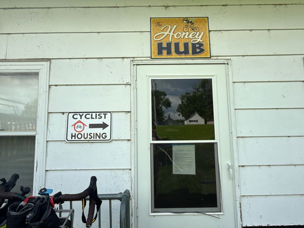 Photograph of a door on a white house with a sign next to it that says “Cyclist Housing” and an arrow pointing to the door and a sign above it that says “The Honey Hub”
