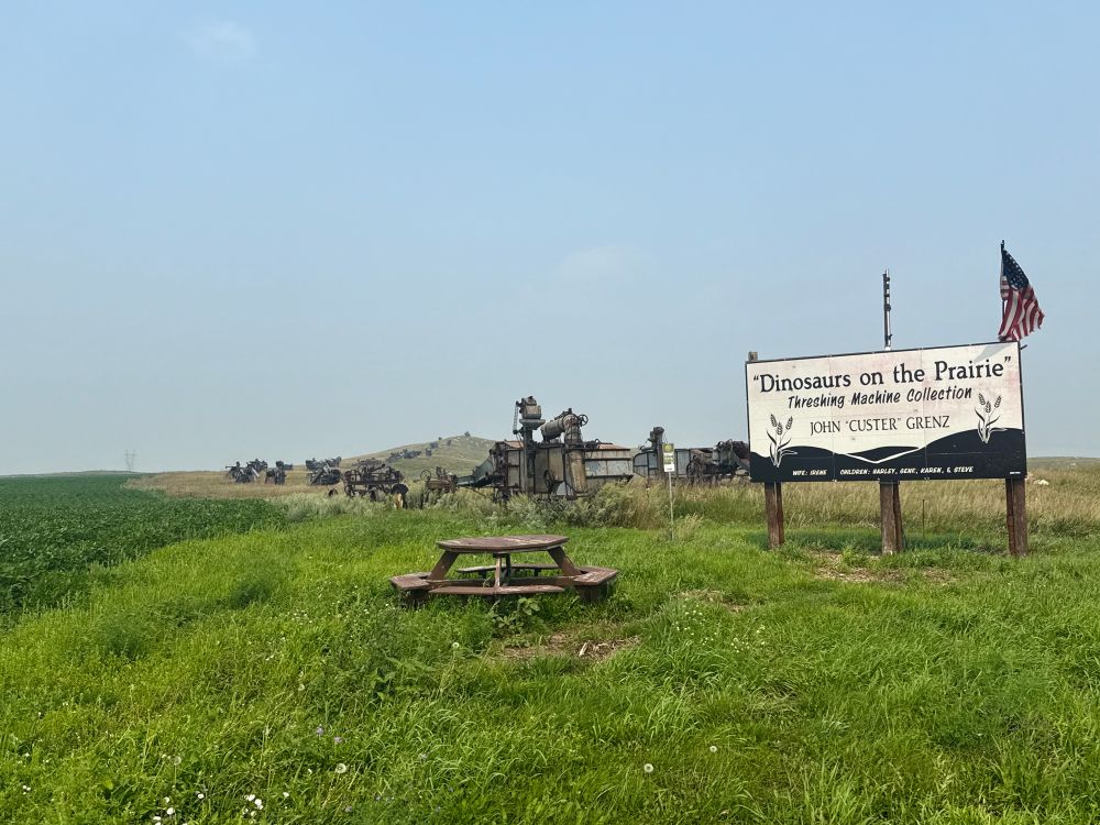 Photograph of a sign saying “Dinosaurs on the Prairie, Threshing Machine Collection” next to a picnic table with a line of derelict threshing machines, presumably, stretching out behind it. 