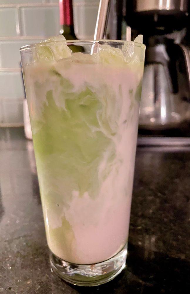 Glass of taro coconut matcha milk tea. Swirls of green and a soft purple inside the glass with ice. There’s a metal straw in the glass. 
