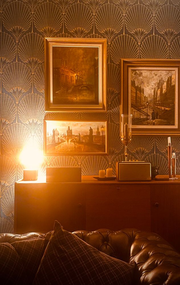 En bild på en vägg. På väggen hänger tre tavlor med stadsmotiv från Amsterdam och Prag. Framför väggen står en skänk i teak, ovanpå skänken står en bordslampa, en humidor, en högtalare och lite ljus. Framför skänken står en chesterfieldfåtölj med en rutig kudde i.