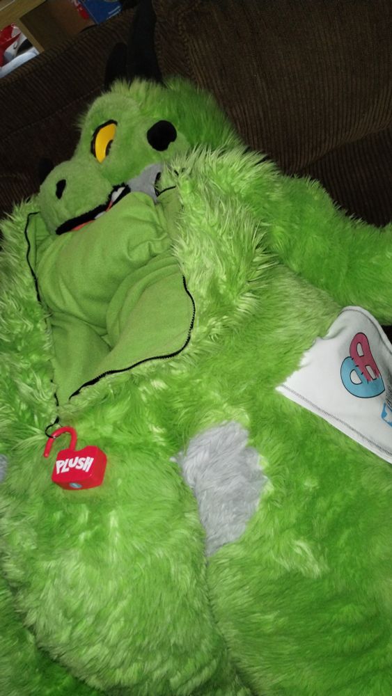 Paradise the Plush dragon laying on a brown couch, empty, zipper open exposing the internal lining. A large plastic lock with the word Plush on it is hanging by the zipper