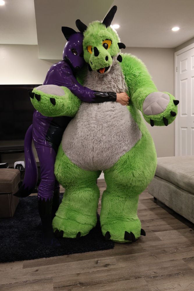 Paradise the plush dragon standing in the middle of a living room. A purple rubber fox is hugging them from the side