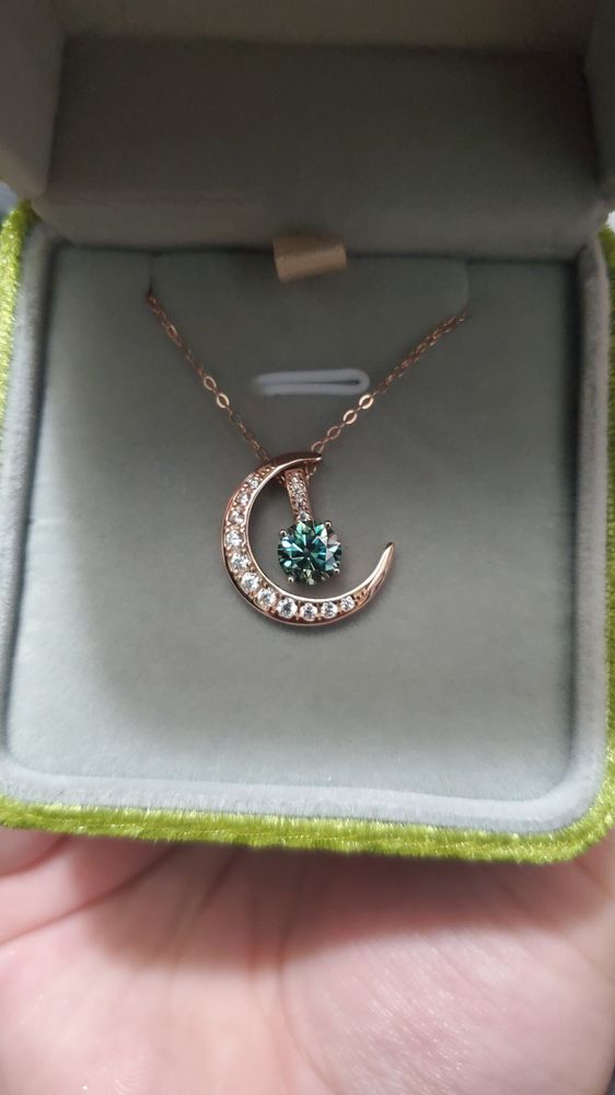 A necklace in a jewelry box. The necklace is made out of rose gold, with a thin rose gold chain. Its pendant is a crescent moon studded with tiny white gems, and a bright green round moissanite stone sits in the center of the crescent. It is attached to the main pendant by a rose gold loop studded with tiny white gems. 