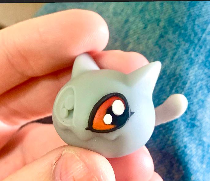  The Digimon “Nyaromon” as a resin 3D print. It looks like a limbless blob with feline features such as ears and a tail. It’s got big, shiny eyes and a zigzag grin. It’s looking to it’s left, kinda mischievous. 

 It’s mostly grey and unpainted, except for its left eye, which I’d spontaneously decided to practice painting on. Two big white highlights, black pupil and outline. There’s no white sclera, it’s just a gradient of orange gradating to red towards the top. 

 I’m pretty proud of it all for a first try!