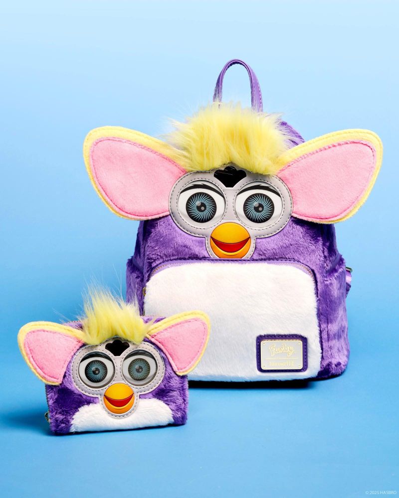 A wallet and backpack, in the design of a purple Furby with white stomach, blue-grey eyes and yellow ears and hair tufts, are presented on a gradient soft blue background. The wallet sits in front of and to the left of the backpack.