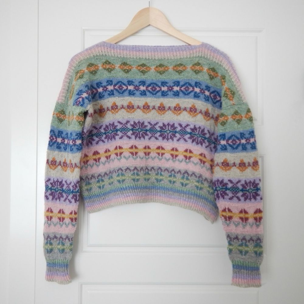A colourful fair isle style hand knit sweater hangs on a coat hanger against a white door. The sweater has colourful striped stranded colourwork sections with pastel shades of green, pink, purple and blue.