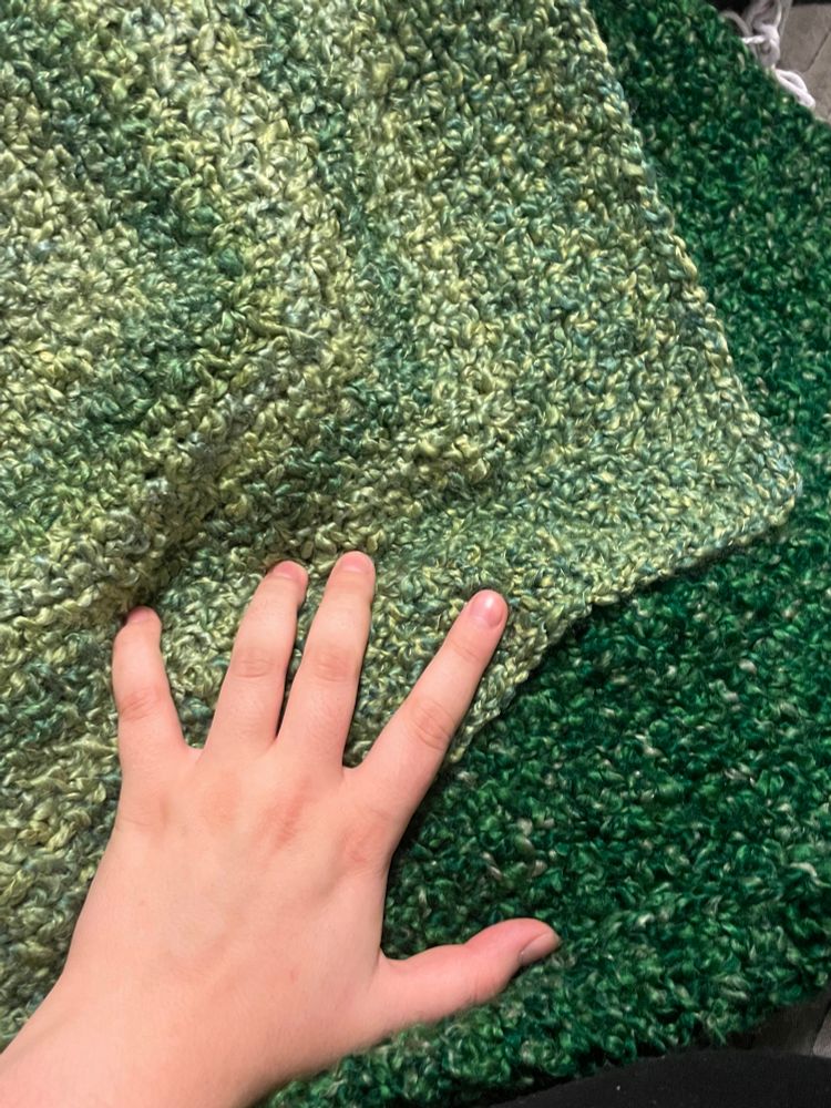 The creator’s hand rests on top of two green shawls. The shawl on top is lighter green with variation and the shawl beneath it is a dark green with speckles.