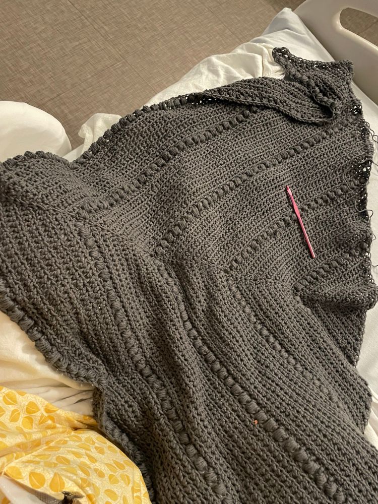 A completed dark grey crochet shawl resting on a hospital bed with a pink hook on top of it. The creator’s yellow gown is visible in the bottom left corner. 