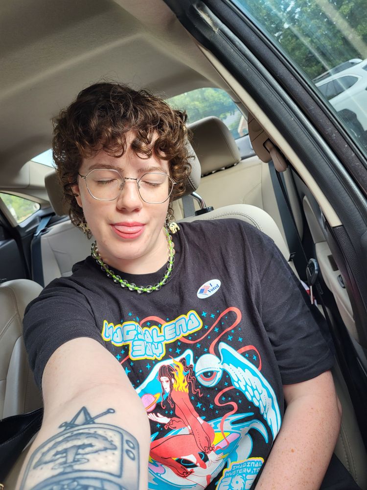 Selfie of me taken in my car! My brown curly hair is in a pixie/mullet with bangs. I'm wearing glasses, a fun green beaded necklace, a Magdalena Bag shirt, and an "I voted" sticker