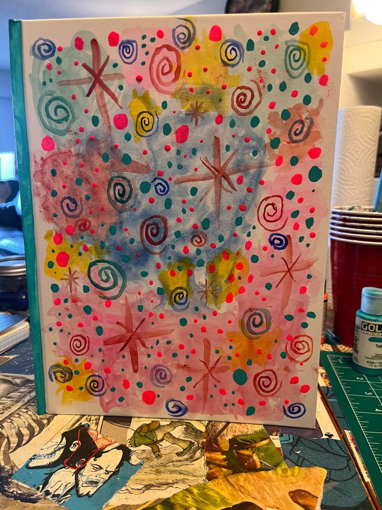 a "letter sized" notebook standing up on a collaged table. it has been painted over with blotchy watercolours ans messy spirals and stars and little teal and flourescent pink dots