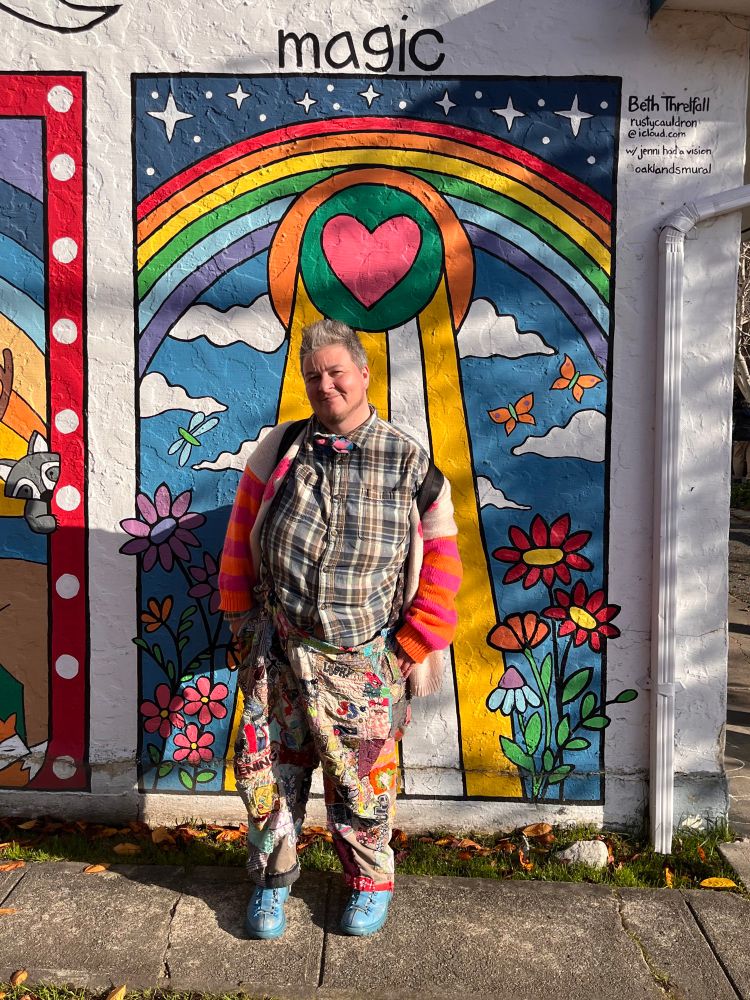 a fat white human wearing patched pants, a cardigan with bright pink and orange sleeves, blue boots, a collared plaid shirt and a blue and pink bowtie standing in front of a mural with a heart beaming down from a rainbow and the word magic on top