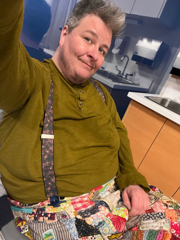 fat white human in a green long sleeved shirt and patched pants sitting in front of a clinical looking sink area