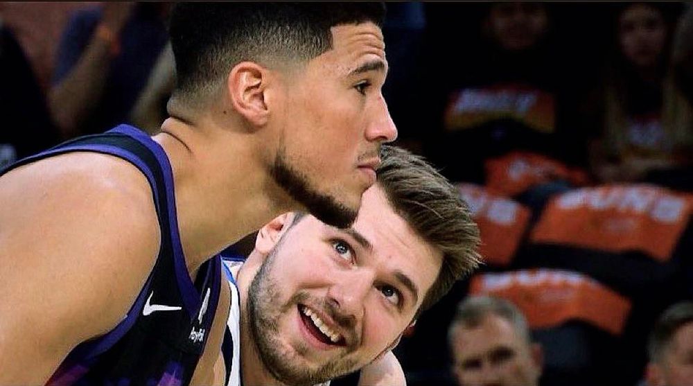 Luka Doncic looking at Devin Booker smiling while Booker is very serious and trying to ignore Luka 