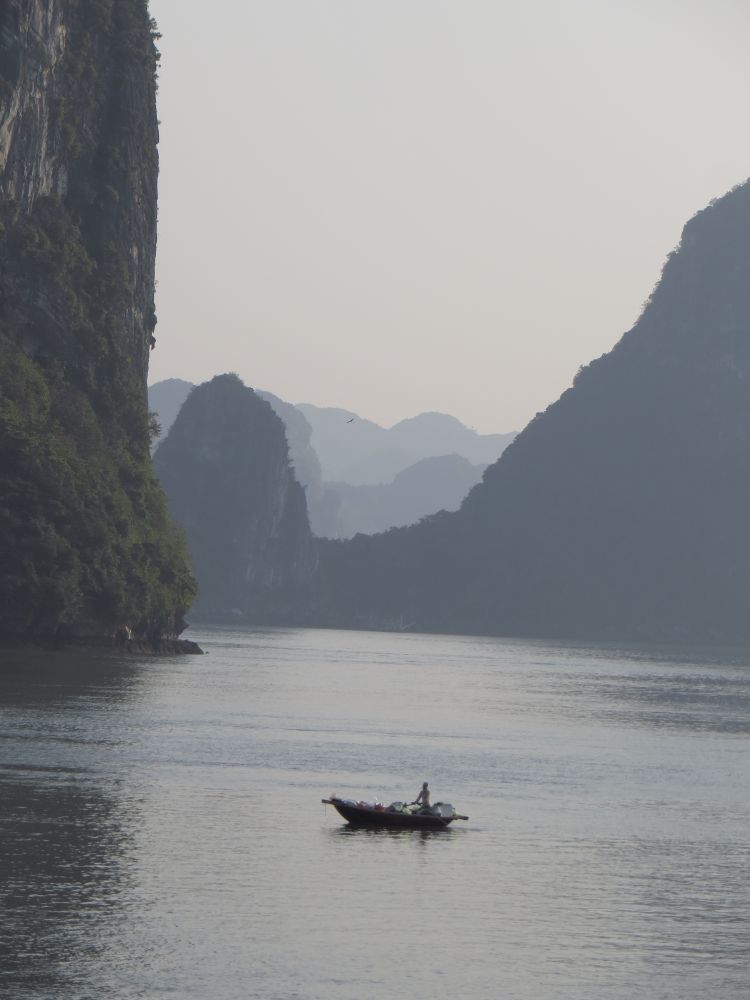 Ein Gewässer. Im Hintergrund steile Felsen, die sich im Dunst verlieren. Im Vordergrund ein Mensch auf einem kleinen Boot. 
