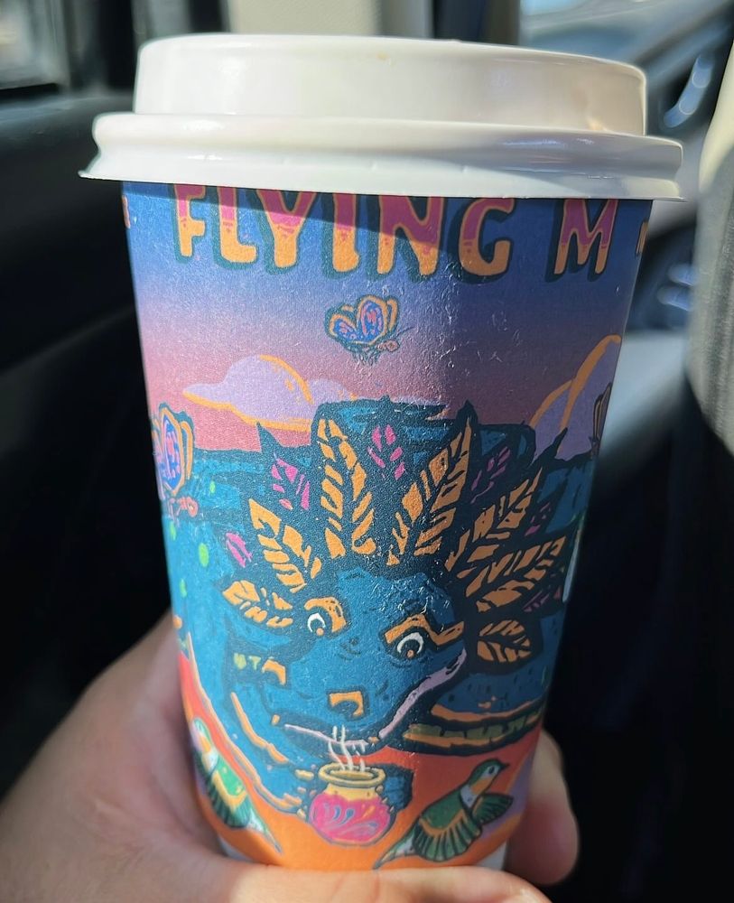 A to go coffee cup featuring art of a blue mythical dragon-like creature with yellow and pink feathers coming out of its head sniffs a steaming pink and yellow cup of coffee. There is a purple and orange sunset happening with a cloud and butterfly on the coffee cup. Orange and yellow all caps letters read “FLYING M” just under the white plastic lid. 
