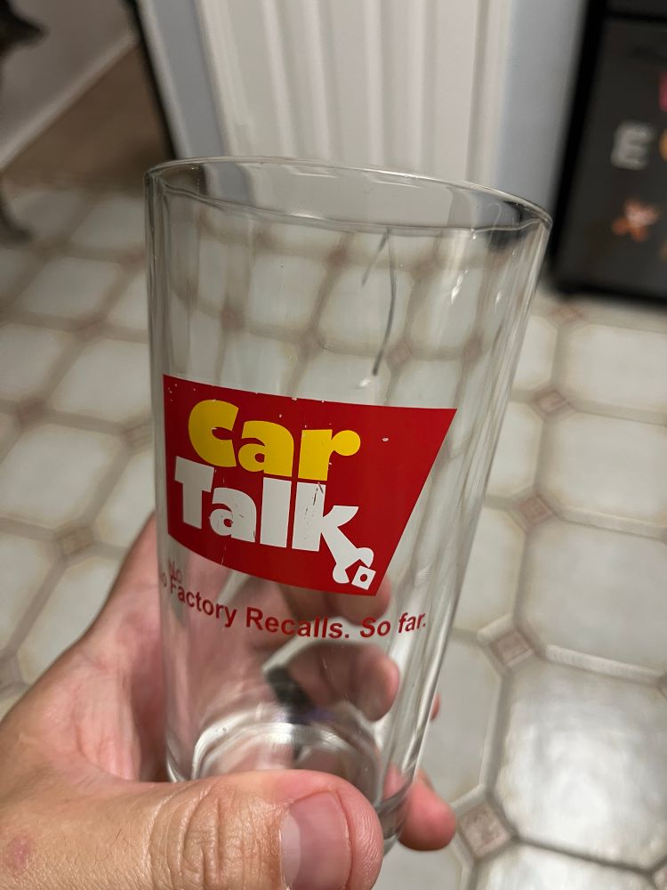 A pint glass with the logo from the former radio show “Car Talk” printed on it is held in someone’s left hand. The logo has the words Car Talk printed in yellow and white respectively over a red background with the words “No Factory Recalls. So far.” printed in red below. At the top of the pint glass, a thin crack runs a couple of inches from the top of the glass down one side. 