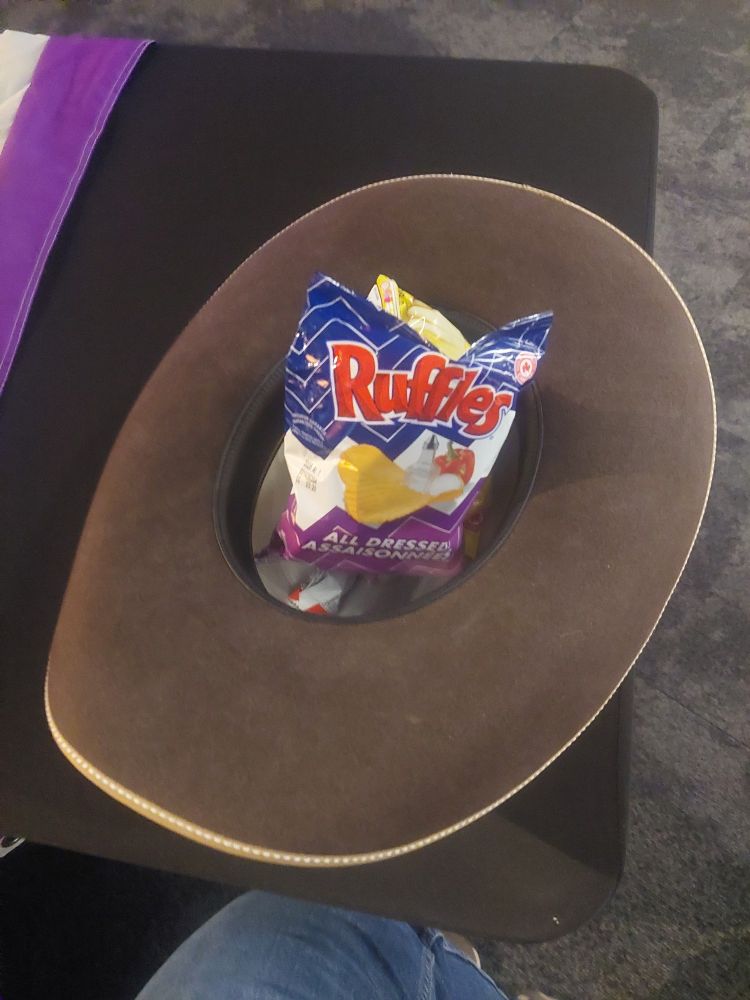 A brown, felt cowboy hat on a black table. There are a few bags of chips in the hat. 