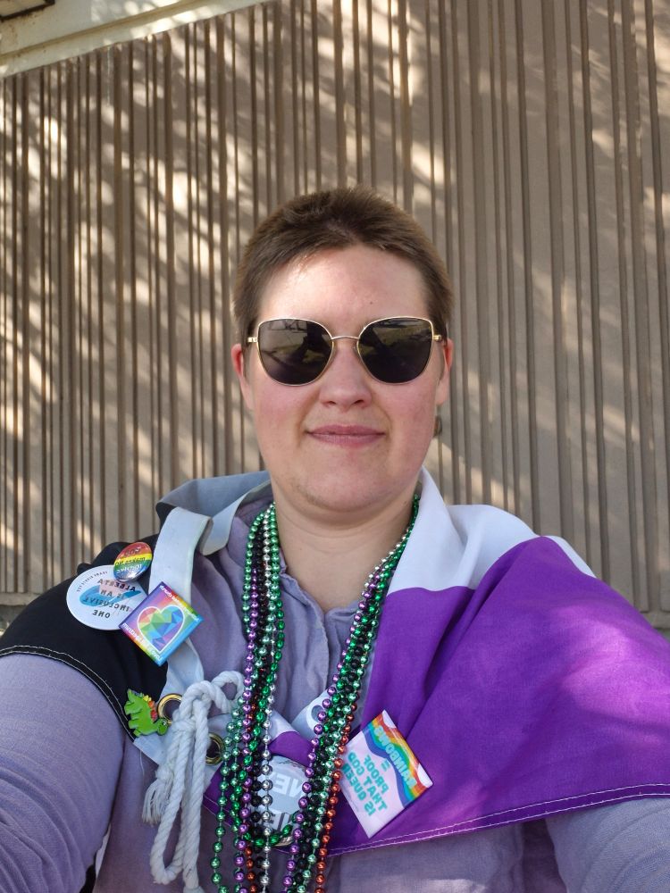 Photo of me in big shades and wearing an asexual flag as a cape. I have tons of needed necklaces and pins