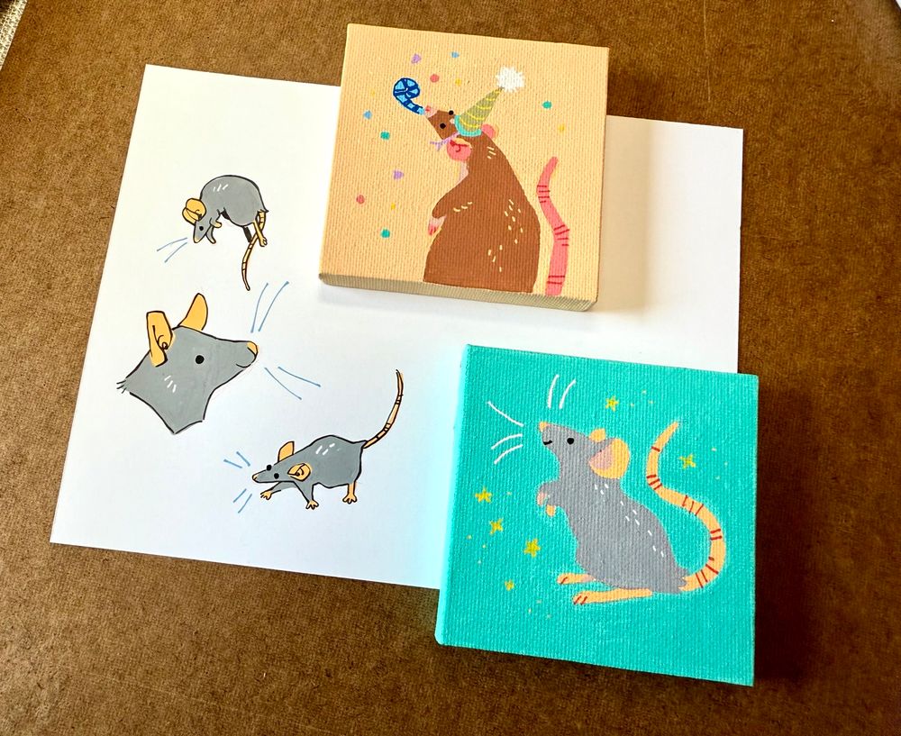 Photo of my work table showing a sketch page and 2 3x3” canvases with little paintings of little rats done in posca, gouache and a brush liner