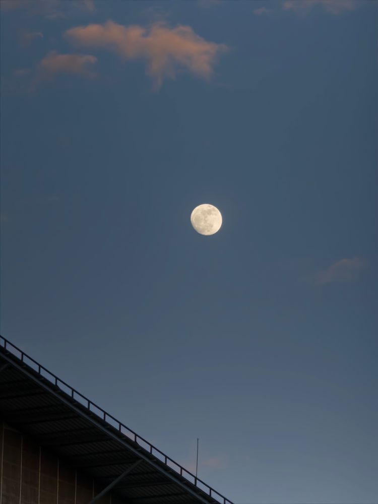 A clear evening sky with a bright, nearly full moon centered above a building with a slanted roof, a few orange-tinged clouds softly floating nearby.