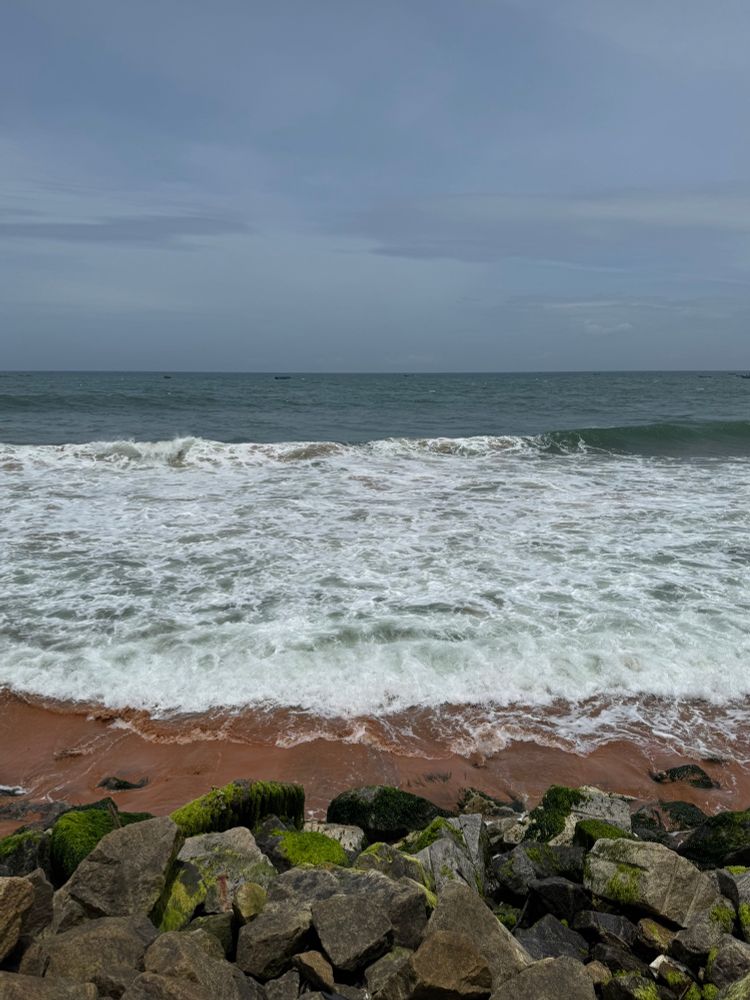 A scenic view of the ocean with waves crashing onto a sandy shore, bordered by large rocks covered in green moss under a cloudy blue sky.
