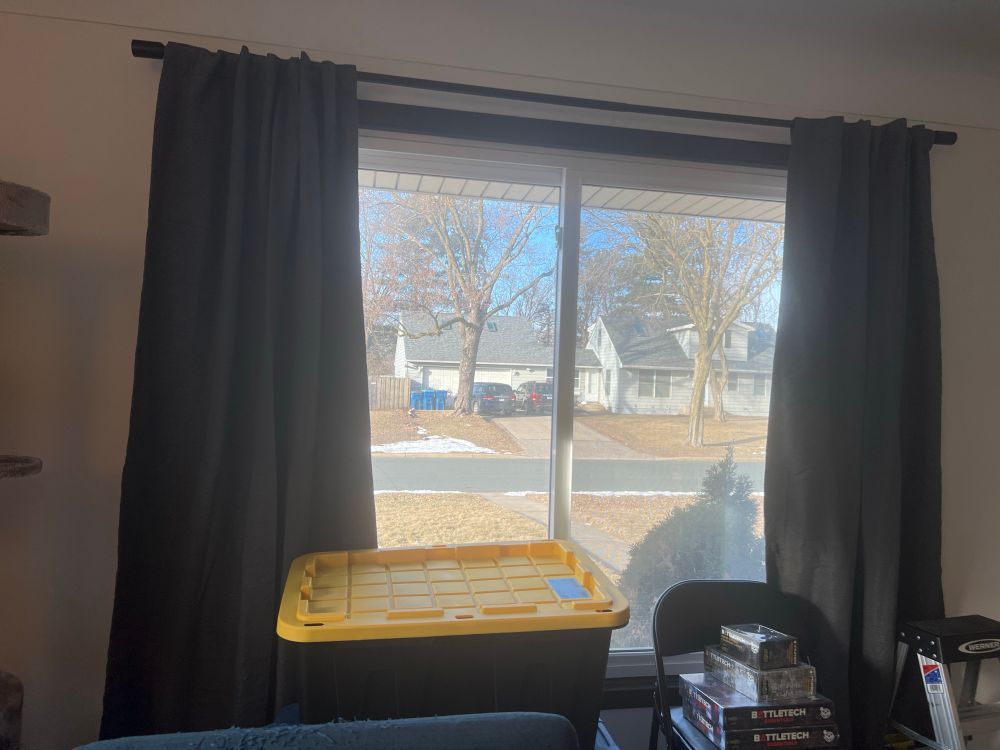 A room with white walls. A large window has a black curtain rod and grey curtains mounted above it, and a few moving boxes can be seen next to a step ladder. 