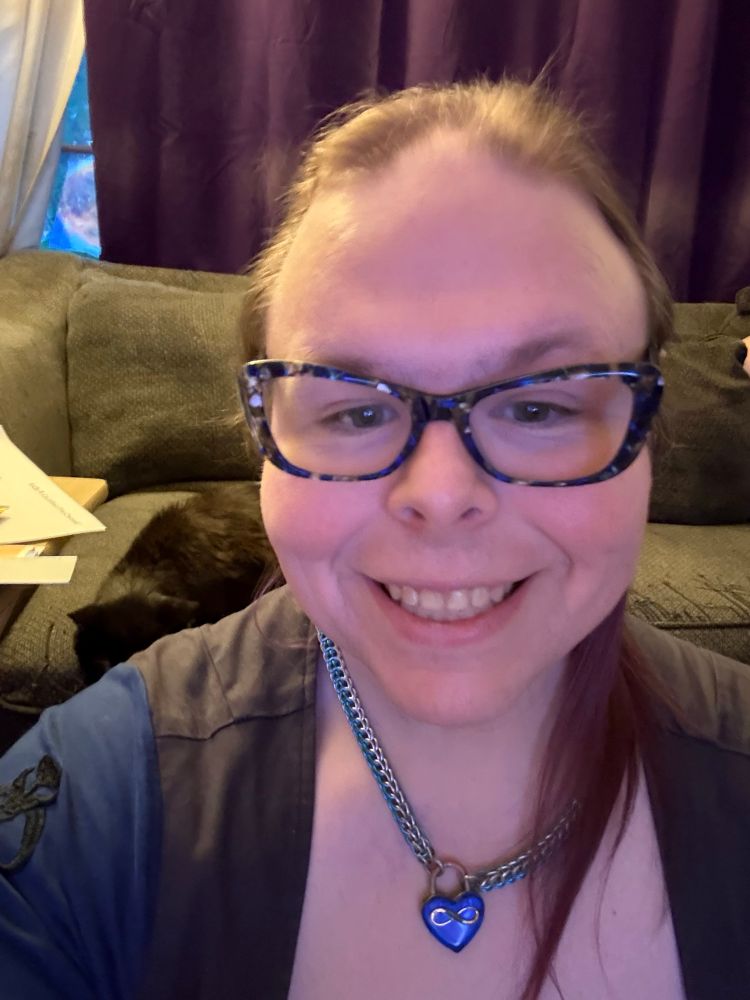 A transgender woman wearing glasses and a day collar smiles at the camera. Her shoulder length hair is a muted purple color. 