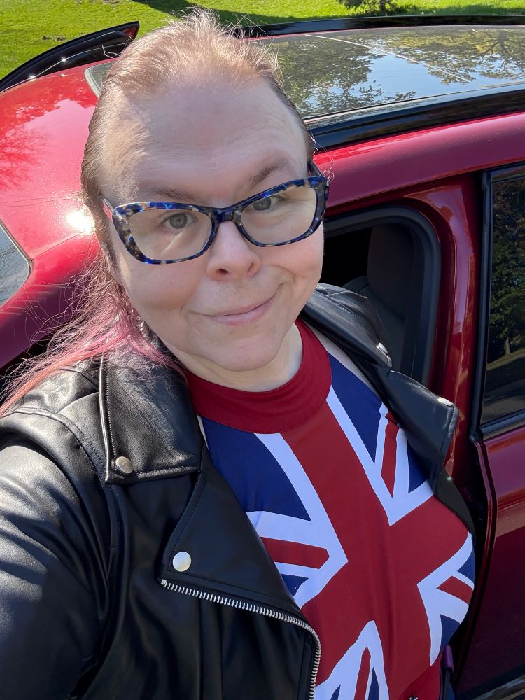 A woman with purple hair and blue glasses smirks at the camera. She is wearing a Captain Carter cosplay with a leather motorcycle jacket. 