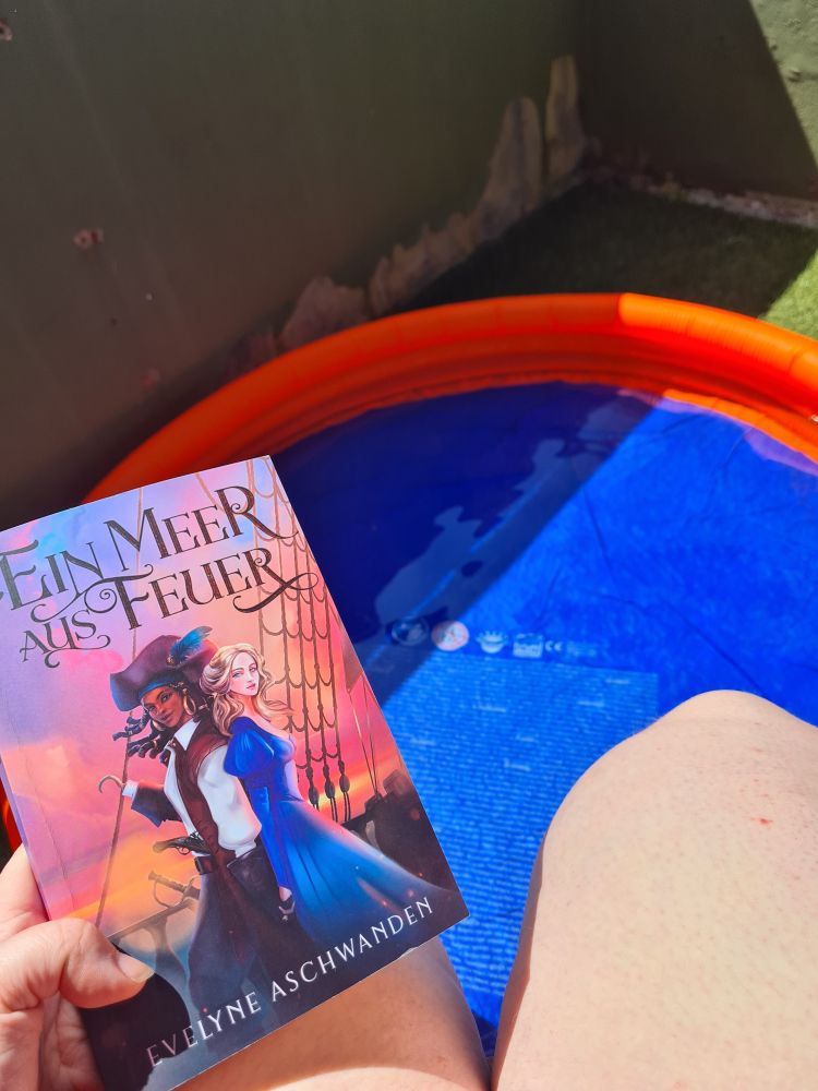 The leg of a white person standin in an orange and blue kiddie pool, holding the book "Ein Meer aus Feuer" 