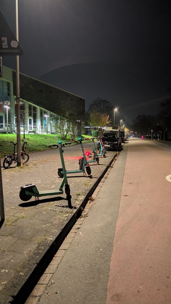 5 eScooter stehen in einer Reihe auf dem Gehweg, auf dem sonst bei jeder Gelegenheit PKW abgestellt werden.

Es ist Abend.