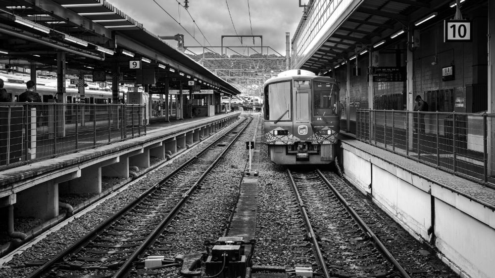 This image is taken from the viewpoint of standing between two railroad platforms while looking down the track. On the right side is a train waiting to be boarded. On either side of the image, people can be seen waiting on the platforms. Image is in monochrome. 