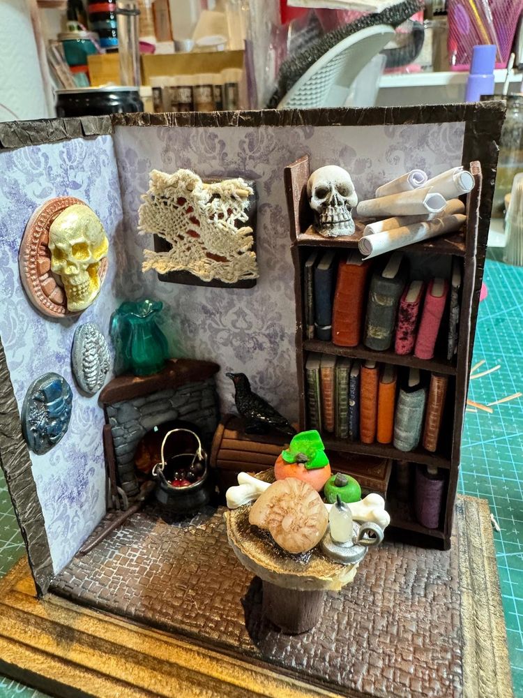 Maqueta proporción 1:12; se ve una habitación de aspecto viejo, chimenea, caldero, decoración de terror, muchos libros.